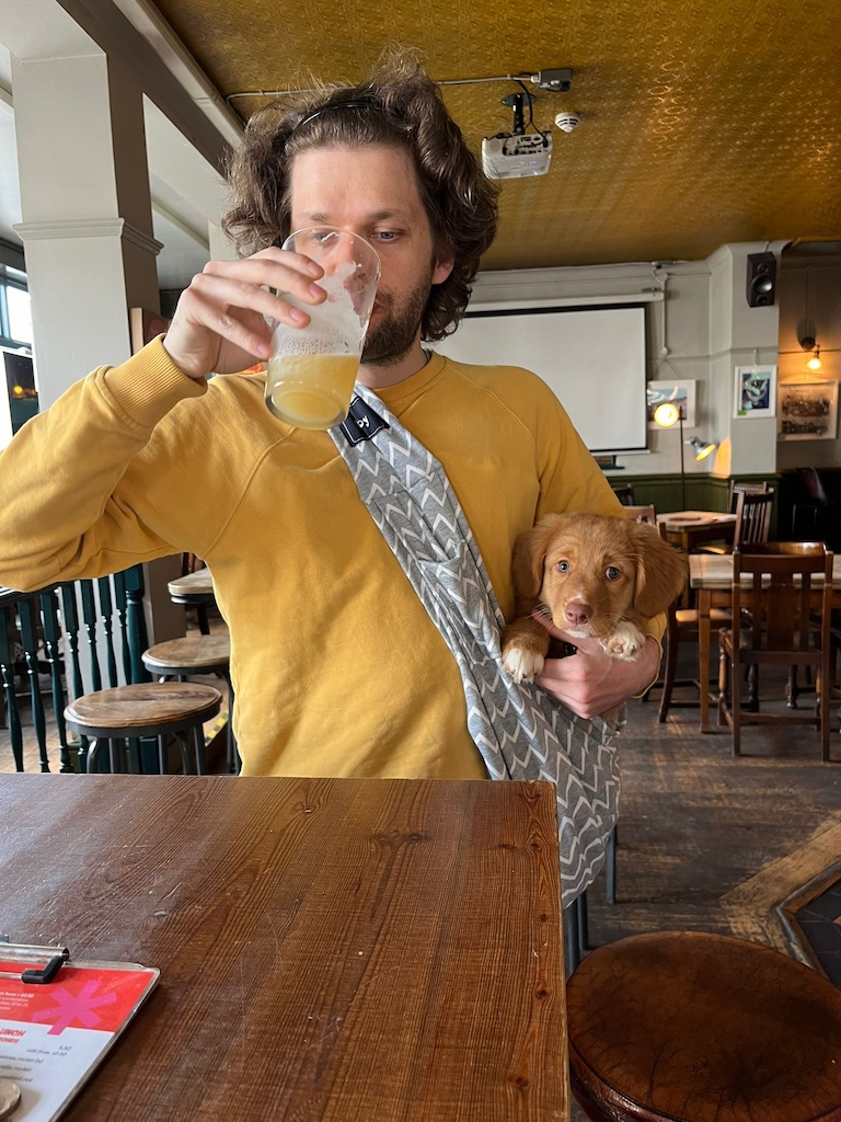 A picture of me holding Rosie, the dog, in a sling whilst drinking a pint in a pub.